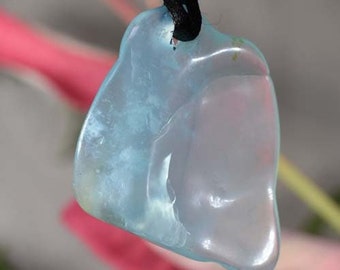 May include: A translucent, light blue pendant with a smooth, organic shape. The pendant is suspended from a black cord, and the background features blurred pink and green elements. The pendant's color shifts from light blue to a pale, almost white hue.