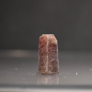 May include: A reddish-brown, rectangular crystal with a faceted top. The crystal has a slightly rough texture and is set against a dark gray background. The crystal is standing upright.