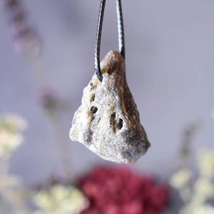 Puede incluir: Un colgante de piedra gris y marr&oacute;n con una superficie rugosa y texturizada cuelga de un cord&oacute;n negro. El colgante tiene forma triangular y tiene un aspecto natural y org&aacute;nico.