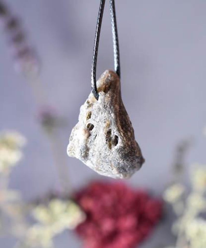 Puede incluir: Un colgante de piedra gris y marr&oacute;n con una superficie rugosa y texturizada cuelga de un cord&oacute;n negro. El colgante tiene forma triangular y tiene un aspecto natural y org&aacute;nico.