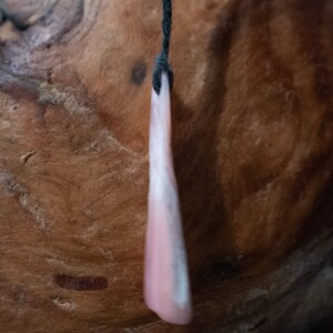 May include: A pink and white pendant hangs from a black cord. The pendant is a long, slender shape with a rounded bottom. The background is a textured, brown wooden surface.