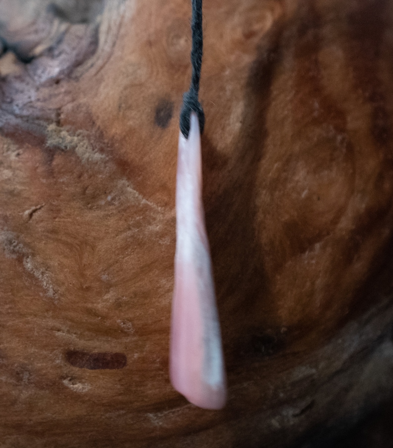 May include: A pink and white pendant hangs from a black cord. The pendant is a long, slender shape with a rounded bottom. The background is a textured, brown wooden surface.