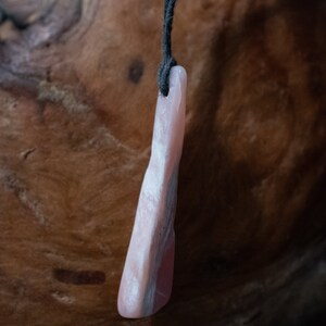 May include: A pink stone pendant with a dark cord. The pendant has a smooth, polished surface with variations in color, including shades of pink and gray. The stone is elongated and irregular in shape, with a slightly curved form.