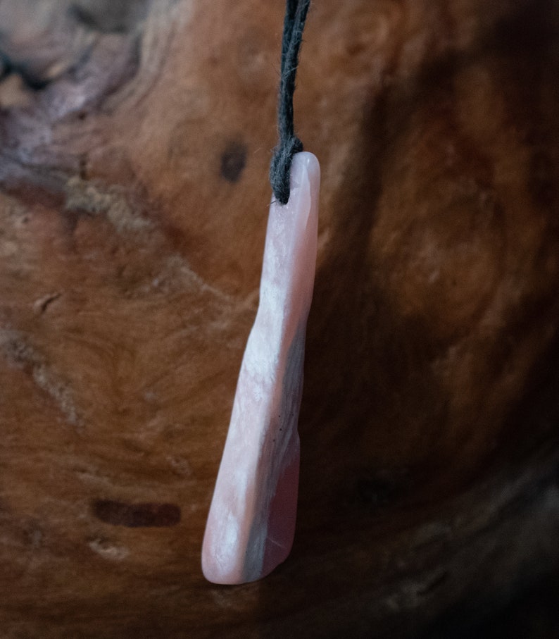 May include: A pink stone pendant with a dark cord. The pendant has a smooth, polished surface with variations in color, including shades of pink and gray. The stone is elongated and irregular in shape, with a slightly curved form.