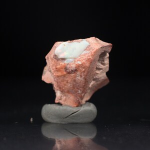 May include: A rough, reddish-brown stone with a large, white opal vein. The opal displays flashes of iridescent color. The stone is mounted on a small, gray, rounded base, set against a black background.