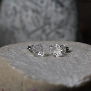 Puede incluir: Un par de pendientes de botón con piedras transparentes de forma irregular y facetadas. Cada piedra está montada en un soporte metálico, mostrando una superficie rugosa y texturizada con múltiples ángulos reflectantes y un aspecto translúcido.