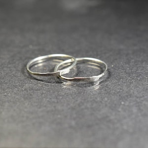 Può includere: Due delicati anelli d'argento sono intrecciati, su uno sfondo scuro. Gli anelli hanno una texture martellata, che cattura la luce e crea sottili riflessi. Il design minimalista suggerisce un'attenzione alla semplicità e all'eleganza.