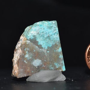 May include: A close-up of a rough-cut stone with a teal-colored surface and a dark brown base. The stone sits on a small, gray, sculpted base. A copper coin is visible in the background.