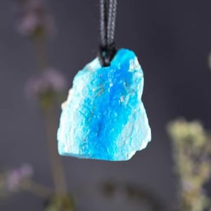 Może przedstawiać: Wisiorek z żywym niebieskim kamieniem szlachetnym zawieszony na czarnym sznurku. Kamień ma szorstką, teksturowaną powierzchnię z ciemniejszymi niebieskimi smugami. Unikatowa biżuteria.