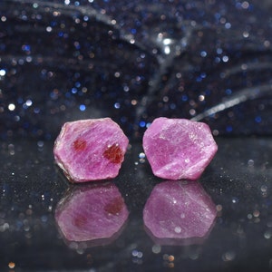 May include: Two rough-cut, hexagonal ruby gemstones. The rubies are a deep pinkish-red color with some darker red inclusions. The gemstones are displayed on a reflective black surface with a dark blue, glittery background.