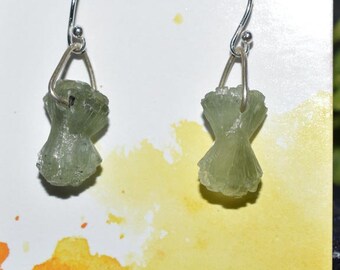 May include: A pair of earrings featuring light green, textured, raw gemstone pendants. The earrings have silver-colored ear wires and a small silver-colored wire connecting the gemstone to the ear wire. The background is white and yellow.