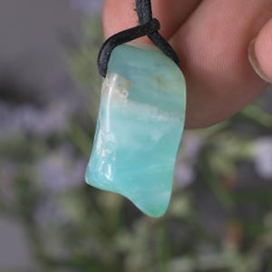 Peut inclure: Pendentif en pierre pr&eacute;cieuse bleu clair avec un cordon noir. La pierre pr&eacute;sente une surface polie avec des nuances de bleu clair et fonc&eacute;. Le pendentif est de forme irr&eacute;guli&egrave;re, sugg&eacute;rant une pierre naturelle.