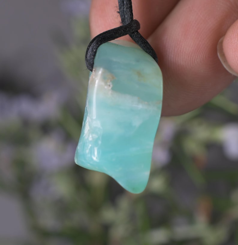 Peut inclure: Pendentif en pierre pr&eacute;cieuse bleu clair avec un cordon noir. La pierre pr&eacute;sente une surface polie avec des nuances de bleu clair et fonc&eacute;. Le pendentif est de forme irr&eacute;guli&egrave;re, sugg&eacute;rant une pierre naturelle.