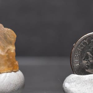 May include: A raw, translucent, amber-colored gemstone sits atop a small white pedestal. A U.S. dime is next to the gemstone for size comparison. The background is a solid, dark gray.