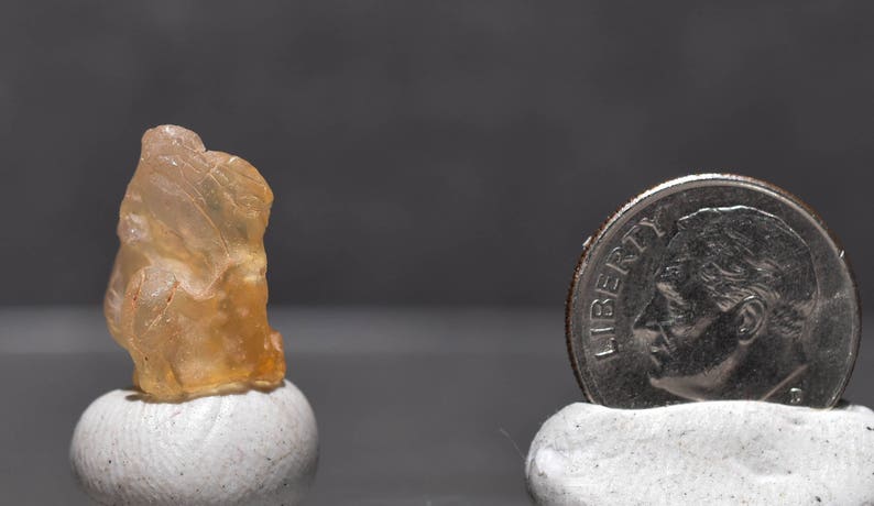 May include: A raw, translucent, amber-colored gemstone sits atop a small white pedestal. A U.S. dime is next to the gemstone for size comparison. The background is a solid, dark gray.