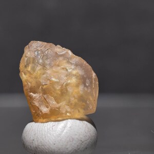 May include: A rough-cut, translucent, amber-colored gemstone. The stone has a textured surface and sits atop a small, white, rounded object. The background is a gradient of gray.