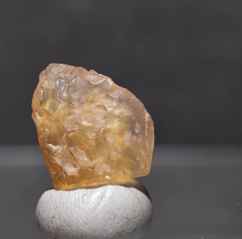 May include: A rough-cut, translucent, amber-colored gemstone. The stone has a textured surface and sits atop a small, white, rounded object. The background is a gradient of gray.