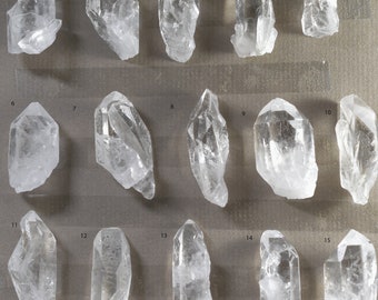 May include: Fifteen clear quartz crystals of varying sizes and shapes, numbered 1 through 15, displayed on a gray surface.