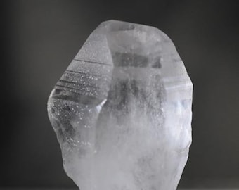 Puede incluir: Un cristal de cuarzo transparente con una punta puntiaguda y una superficie rugosa y texturizada. El cristal está sobre una superficie gris.