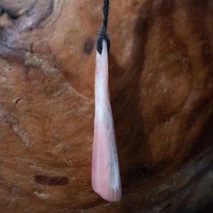 May include: A pink and white pendant hangs from a black cord. The pendant is a smooth, elongated shape with a gradient of pink to white. The background is a textured, brown wooden surface.