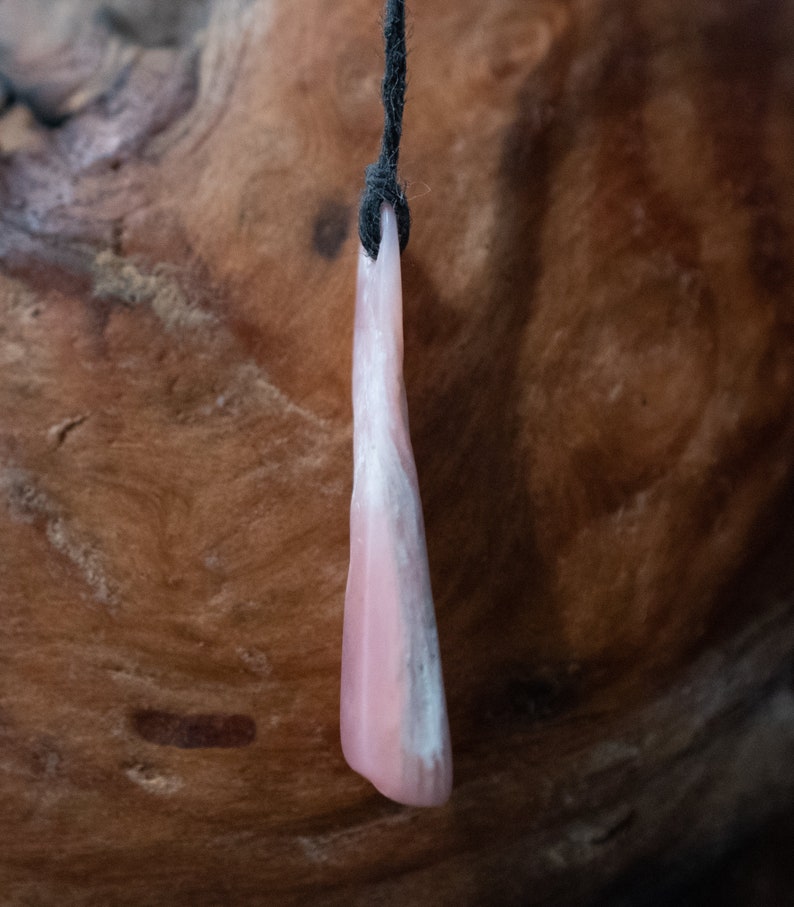 May include: A pink and white pendant hangs from a black cord. The pendant is a smooth, elongated shape with a gradient of pink to white. The background is a textured, brown wooden surface.