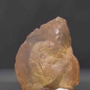 May include: A translucent, amber-colored gemstone with a rough, irregular surface. The stone has internal fractures and inclusions, and is resting on a white surface against a gray background. The overall appearance is natural and organic.
