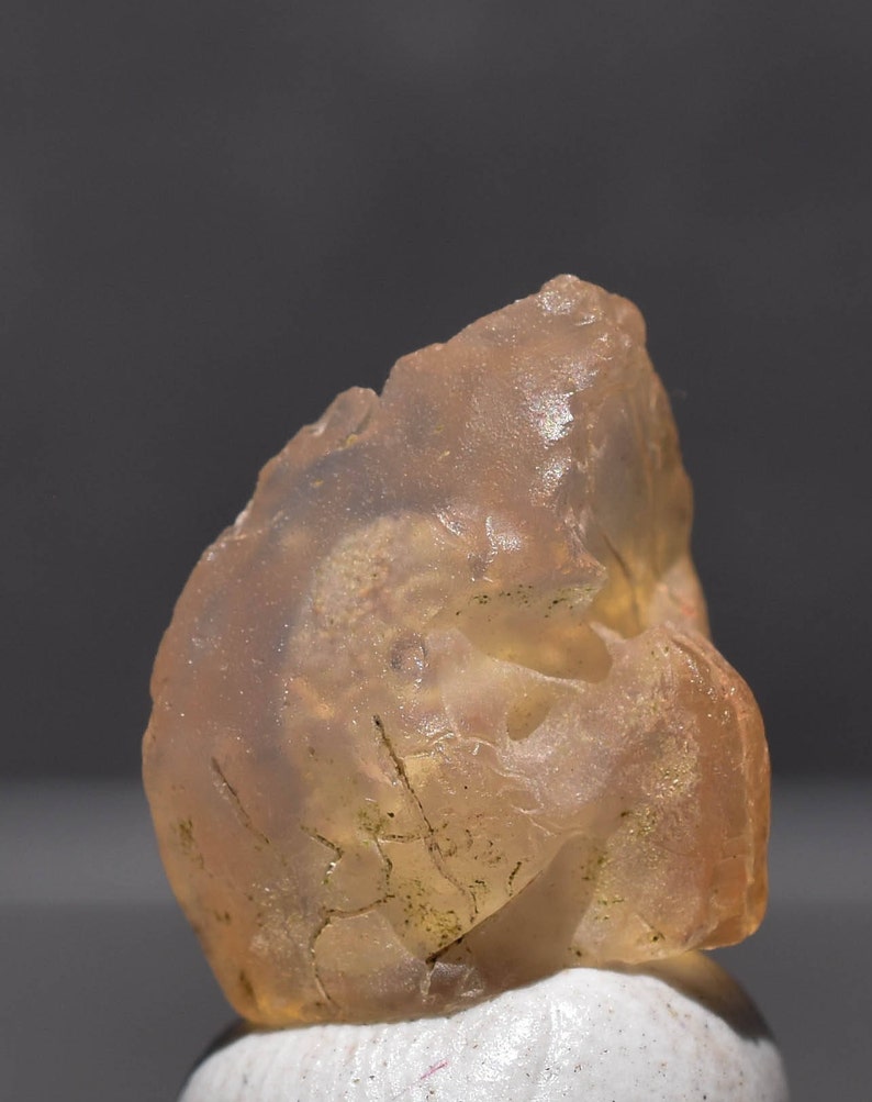 May include: A translucent, amber-colored gemstone with a rough, irregular surface. The stone has internal fractures and inclusions, and is resting on a white surface against a gray background. The overall appearance is natural and organic.