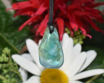 May include: A teardrop-shaped pendant with a teal and blue color scheme, suspended from a black cord. The pendant is held against a backdrop of red flowers and a white daisy with a yellow center.