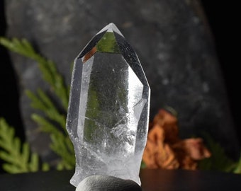 May include: A clear quartz crystal point with a sharp tip and multiple facets. The crystal is sitting on a white surface.