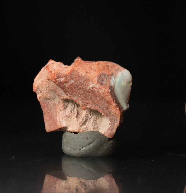 May include: A rough, reddish-brown rock with a section of iridescent opal. The opal displays flashes of green and blue. The rock sits on a small, gray base against a black background. This is a natural gemstone.
