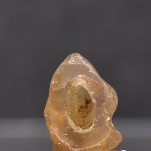May include: A translucent, amber-colored gemstone with a central hole. The stone has a smooth, polished appearance and a natural, organic shape. The gemstone is set against a neutral background, highlighting its color and texture.