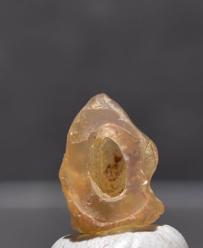 May include: A translucent, amber-colored gemstone with a central hole. The stone has a smooth, polished appearance and a natural, organic shape. The gemstone is set against a neutral background, highlighting its color and texture.