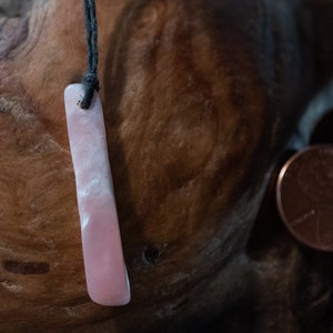 May include: A rectangular, pink pendant with a black cord. The pendant has a smooth, polished surface with white marbling. A penny is next to the pendant. The pendant is likely a necklace.