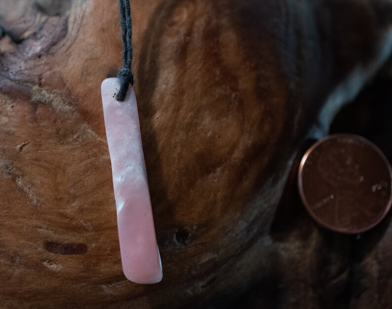 May include: A rectangular, pink pendant with a black cord. The pendant has a smooth, polished surface with white marbling. A penny is next to the pendant. The pendant is likely a necklace.