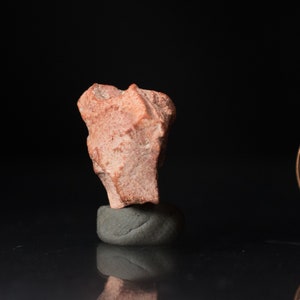 May include: A rough, reddish-orange rock specimen, possibly a mineral or gemstone, is displayed on a small, gray, rounded base. The rock has an irregular shape and texture, with a dark background.
