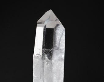 May include: A clear quartz crystal point, approximately 2 inches tall, resting on a gray stone. A copper penny is in the background for size comparison.