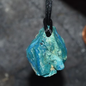 Może przedstawiać: Zbliżenie wisiorka z surowo ciętym, turkusowym kamieniem. Kamień zawieszony jest na czarnym sznurku, ukazując swoją naturalną, teksturowaną powierzchnię. Kolor wisiorka waha się od głębokiego turkusu do jaśniejszego turkusu.