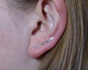 Puede incluir: Un pendiente de plata tipo ear cuff se muestra en una oreja, con cabello rubio visible. El pendiente es una barra fina y recta que se curva para adaptarse a la oreja. El pendiente es de un material metálico brillante, de diseño sencillo y moderno.
