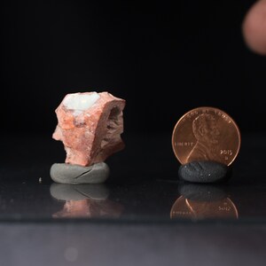 May include: A small, rough-cut, reddish-brown stone with a white, iridescent patch, displayed next to a U.S. penny. The stone sits on a gray, rounded base. The penny is dated 2015 and rests on a black base.