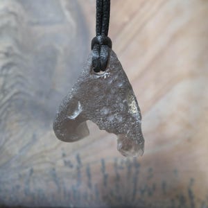 May include: A gray, irregularly shaped pendant made of a translucent material, suspended from a black cord. The pendant has a unique, organic form with a hole near the top and a curved indentation on one side. The background is a blurred, textured surface.