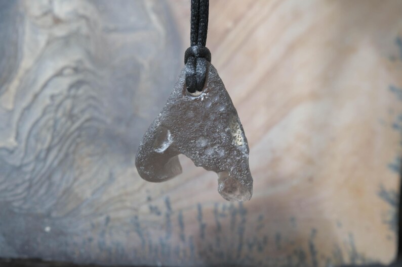 May include: A gray, irregularly shaped pendant made of a translucent material, suspended from a black cord. The pendant has a unique, organic form with a hole near the top and a curved indentation on one side. The background is a blurred, textured surface.