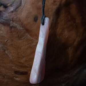 May include: A pink stone pendant with a dark cord. The pendant is a light pink color with darker pink and gray streaks. The stone is rectangular with rounded edges. The cord is black and attached to the top of the pendant.