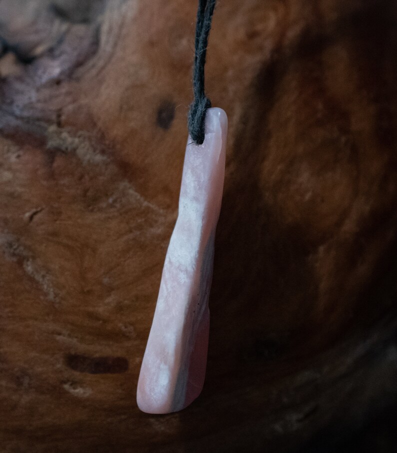May include: A pink stone pendant with a dark cord. The pendant is a light pink color with darker pink and gray streaks. The stone is rectangular with rounded edges. The cord is black and attached to the top of the pendant.