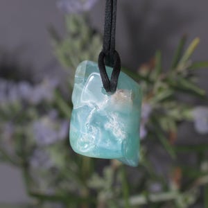 Peut inclure: Un pendentif en pierre pr&eacute;cieuse bleu clair avec un cordon noir. La pierre a un aspect nuageux et marbr&eacute; avec des teintes blanches et bleu clair. Le pendentif est suspendu sur un fond flou de feuillage vert et violet.