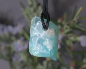 May include: A light blue gemstone pendant with a black cord. The stone has a cloudy, marbled appearance with white and light blue hues. The pendant is suspended against a blurred background of green and purple foliage.
