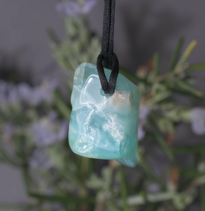 Peut inclure: Un pendentif en pierre pr&eacute;cieuse bleu clair avec un cordon noir. La pierre a un aspect nuageux et marbr&eacute; avec des teintes blanches et bleu clair. Le pendentif est suspendu sur un fond flou de feuillage vert et violet.