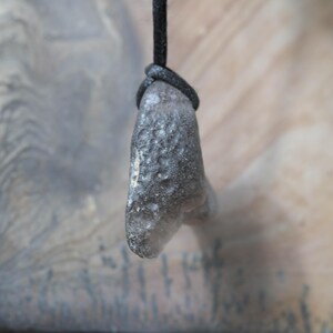 May include: A gray, textured pendant with a dark cord. The pendant has a rounded shape and a rough surface. The cord is knotted at the top, securing the pendant. The background is a blurred, neutral tone.