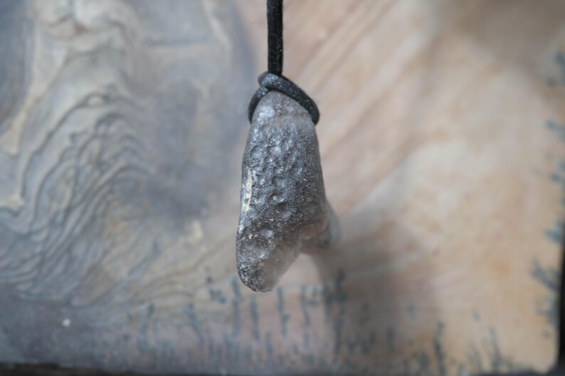 May include: A gray, textured pendant with a dark cord. The pendant has a rounded shape and a rough surface. The cord is knotted at the top, securing the pendant. The background is a blurred, neutral tone.