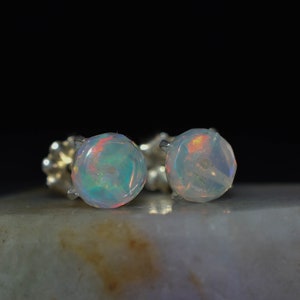 May include: A pair of silver stud earrings with round opal gemstones. The opals are iridescent with shades of blue, green, and pink.