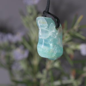 Peut inclure: Un pendentif translucide bleu clair avec un cordon noir. La pierre a un aspect lisse et poli avec des veines blanches et vert clair. Le pendentif est suspendu sur un fond flou de feuillage vert et de fleurs violettes.
